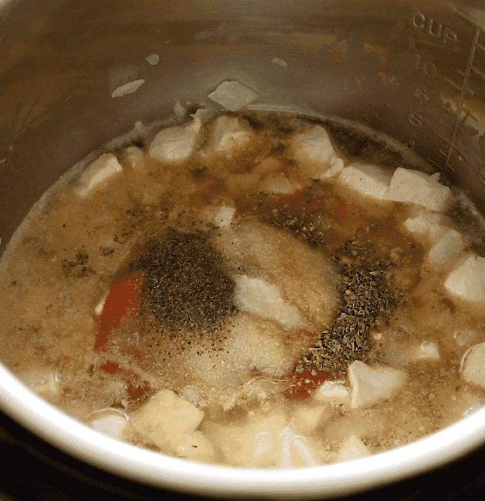 Cubed chicken cooks in a pot with broth, visible seasonings like ground pepper, salt, and herbs floating on top. Measurement markings are on the pots side above the simmering mixture.