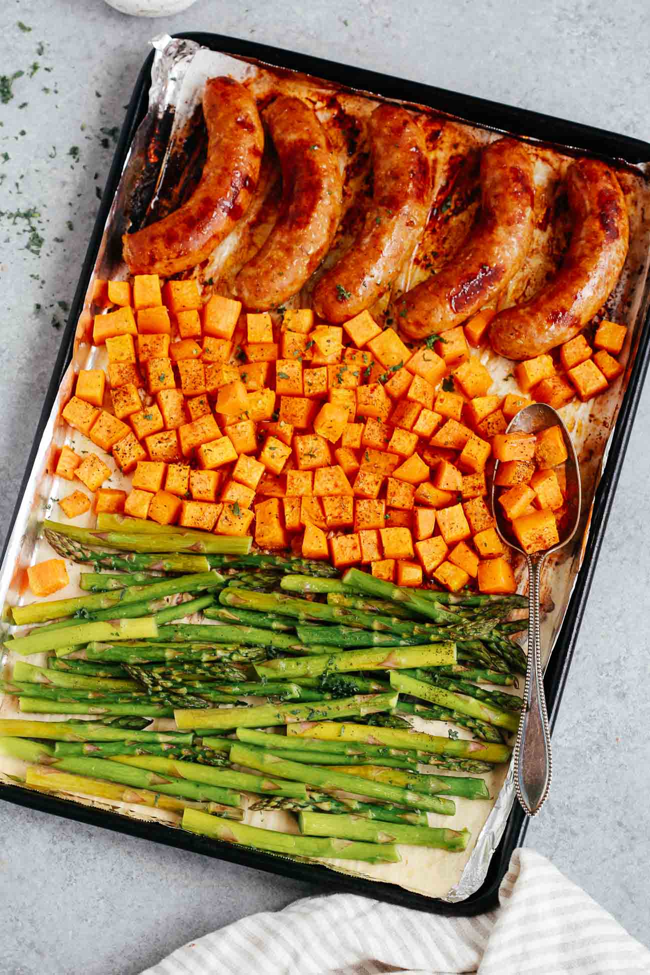 Overhead view of a sheet pan containing cooked sausage, sweet potatoes, and asparagus.