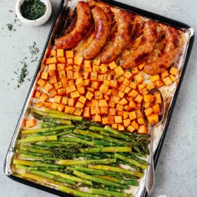 Overhead view of a sheet pan containing cooked sausage, sweet potatoes, and asparagus.