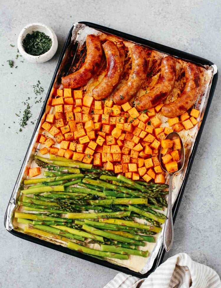 Overhead view of a sheet pan containing cooked sausage, sweet potatoes, and asparagus.