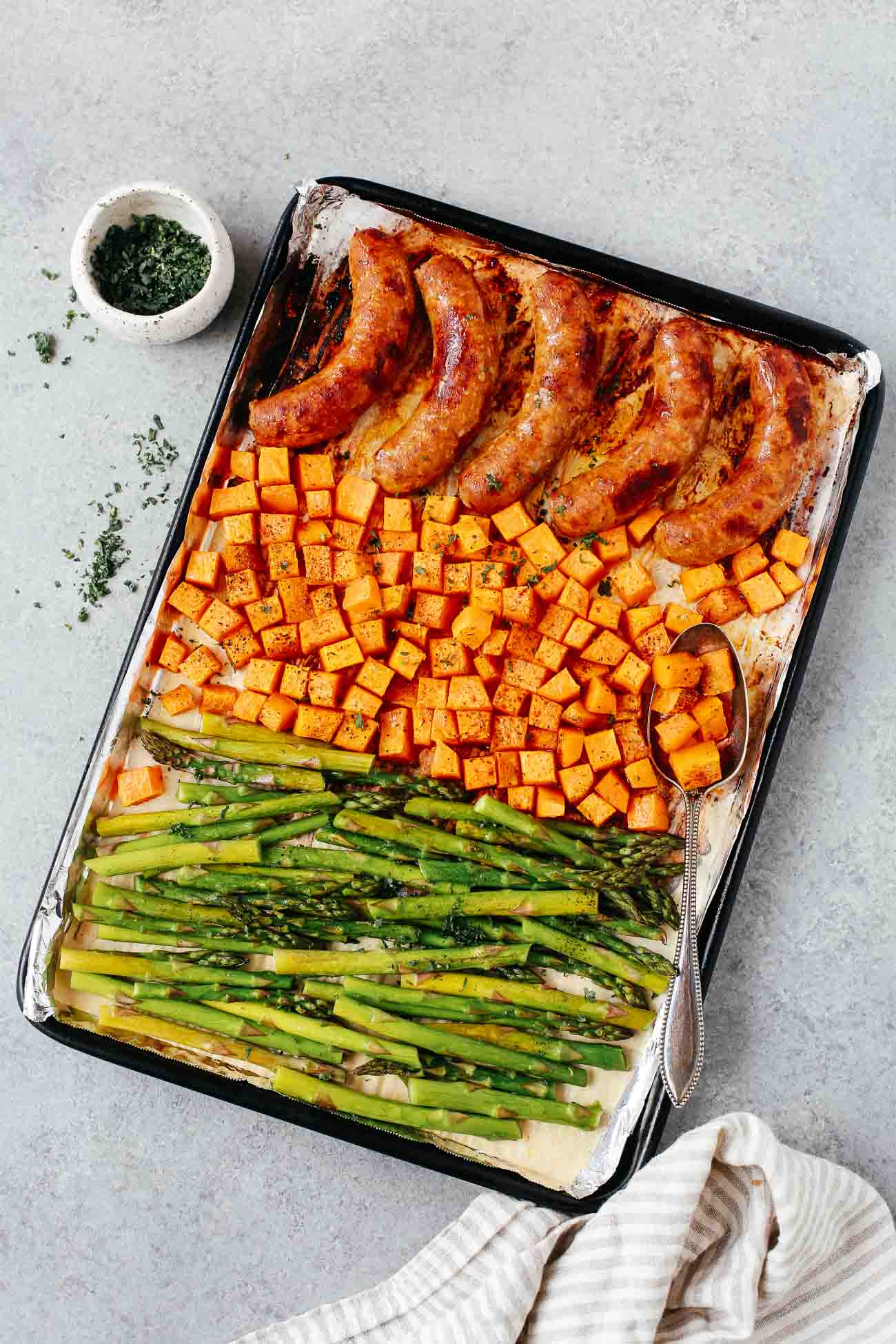 Overhead view of a sheet pan containing cooked sausage, sweet potatoes, and asparagus.