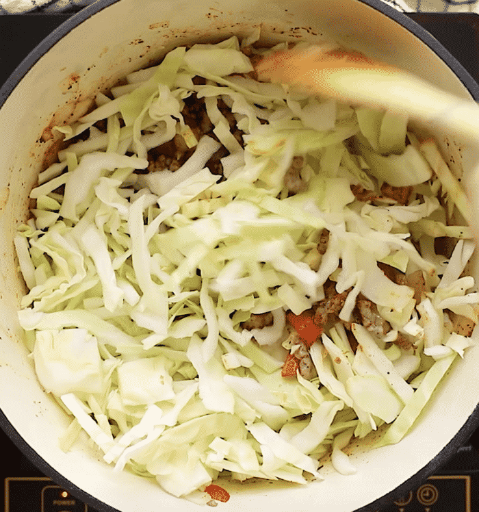 Chopped cabbage is being stirred into a large pot with browned ground meat and vegetables on a stovetop.