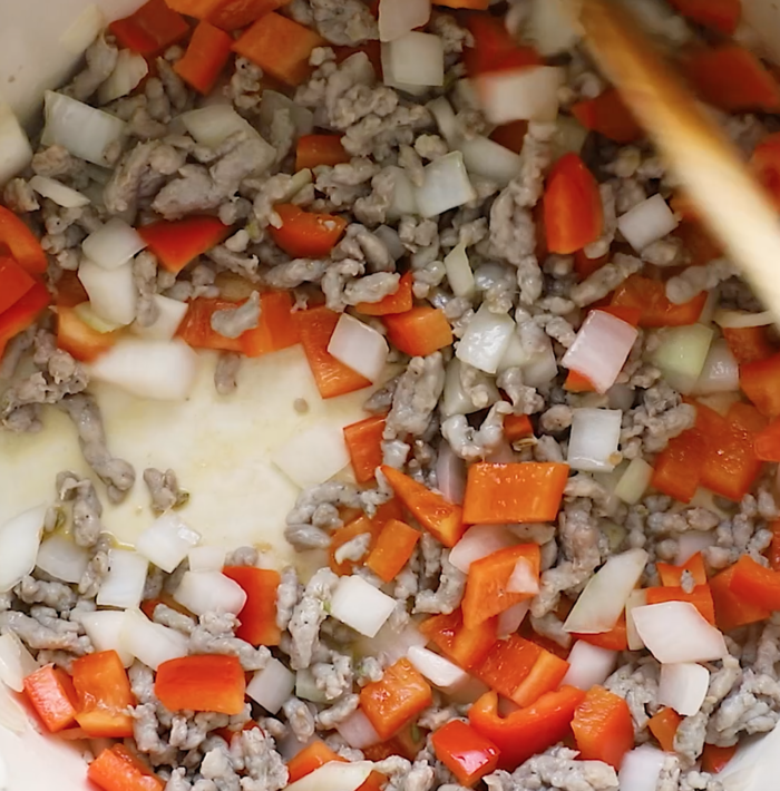 Ground meat, chopped red bell peppers, and diced onions are being sautéed together in a pan, with a wooden spoon stirring the mixture.