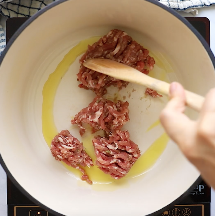 Ground meat being browned in a pot with oil on a stovetop, stirred with a wooden spoon by a hand.