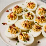 A plate of Easy Deviled Eggs, topped with crumbled bacon and chopped chives, sits on a white surface. A bowl of herbs and a striped napkin are partially visible in the background.