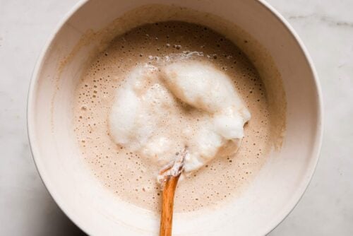 A white mixing bowl contains a frothy, light brown batter for peanut butter waffles, with fluffy egg whites being gently folded in using a wooden spoon. The bowl sits on a light-colored surface.