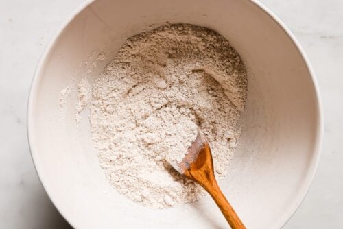 A white mixing bowl filled with dry flour mixture for peanut butter waffles is being stirred by a wooden spoon on a light-colored surface.
