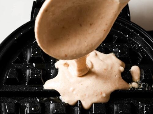 A close-up of peanut butter waffle batter being poured from a spoon onto a black waffle iron, ready to be cooked.