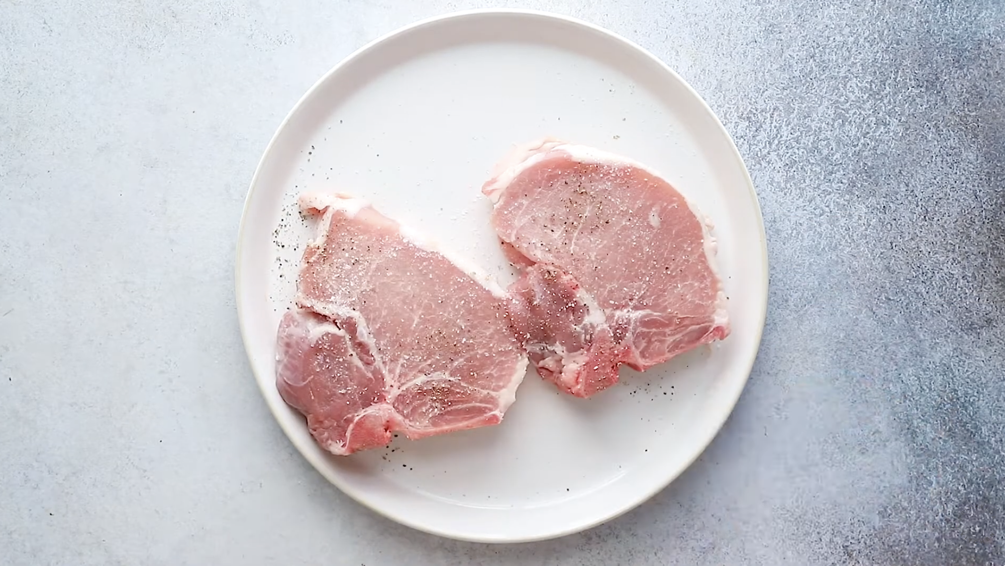 Two raw, seasoned pork chops sit on a round white plate against a light gray background. Sprinkled with salt and pepper, these chops are ready to be transformed into flavorful baked pork chops.