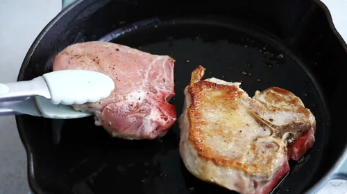 A close-up of two pork chops in a black skillet; one chop is raw and being held with tongs, while the other is seared and browned on one side, perfect for making baked pork chops.