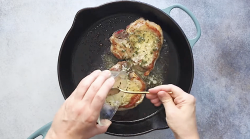 A person pours liquid from a small glass onto two seared baked pork chops cooking in a black cast iron skillet, with herbs and butter visible on the meat.