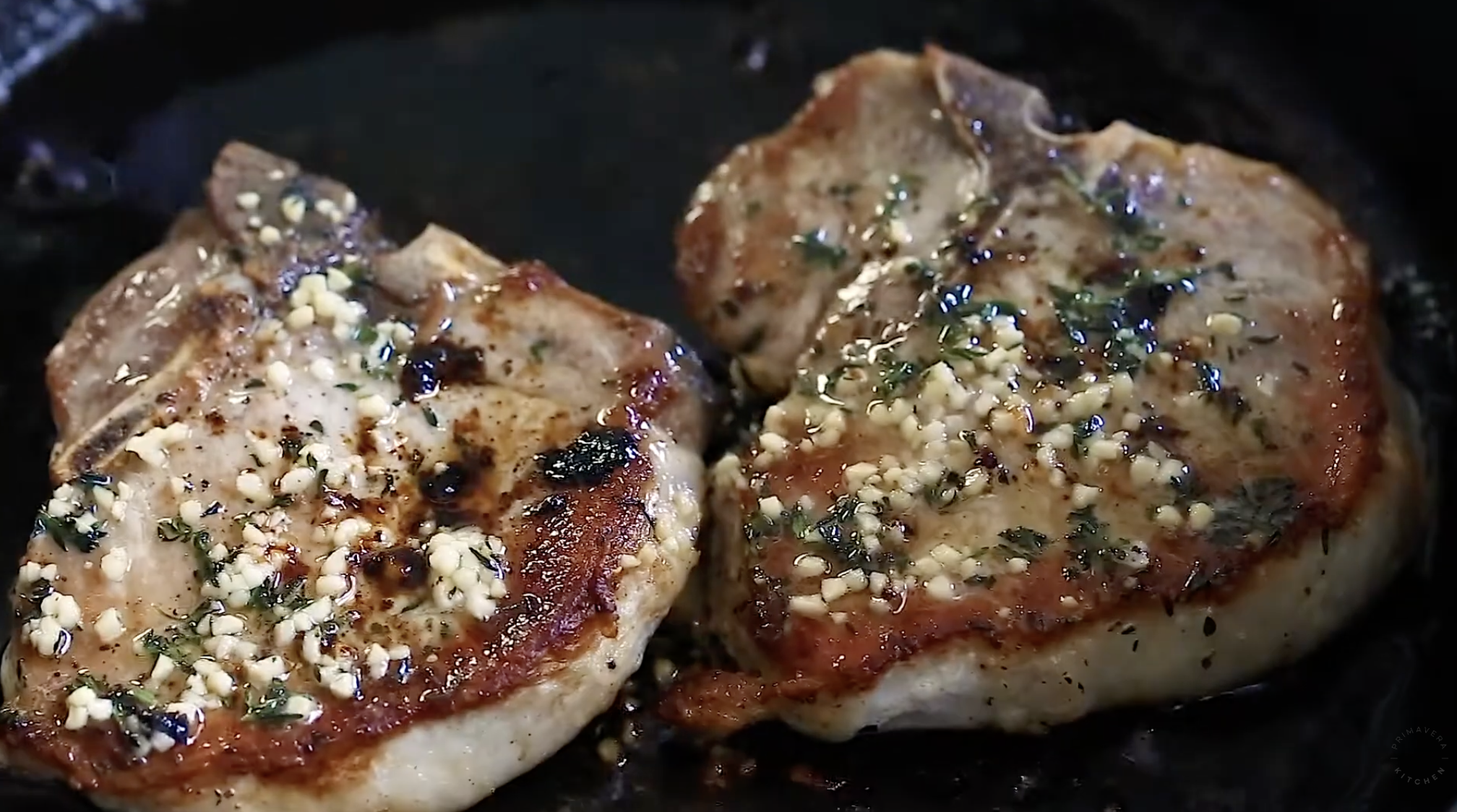 Two bone-in pork chops cooking in a pan, topped with chopped garlic and herbs. These Baked Pork Chops have a golden-brown sear on the edges, with savory juices visible in the pan.