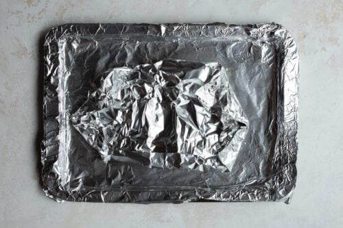 A rectangular baking tray covered with crinkled aluminum foil, holding a smaller, foil-wrapped Rainbow Trout Recipe in the center, sits on a light-colored surface.