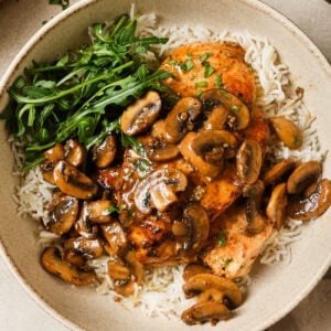 A bowl of white rice topped with flavorful chicken and mushroom, fresh arugula, and grilled chicken breasts, garnished with chopped parsley.