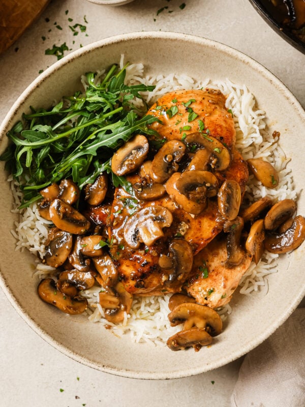 A bowl of white rice topped with flavorful chicken and mushroom, fresh arugula, and grilled chicken breasts, garnished with chopped parsley.