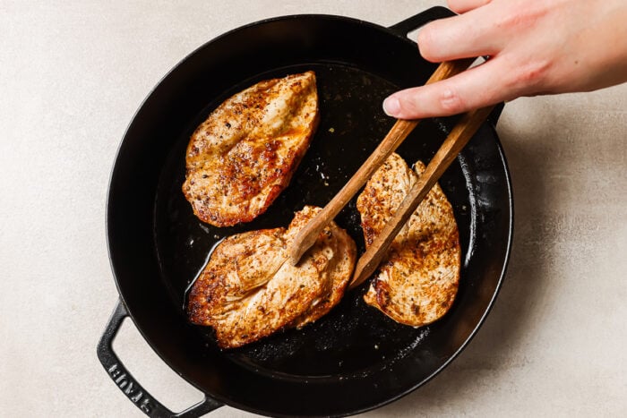A hand uses tongs to flip seasoned, cooked chicken and mushroom breasts in a black skillet on a light-colored surface.