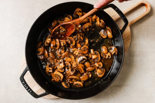 A hand stirs saut&eacute;ed sliced chicken and mushroom in a dark skillet with a wooden spoon; the pan rests on a wooden cutting board atop a light countertop.