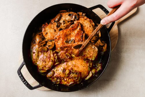 A hand stirs chicken and mushroom in a black skillet on a wooden board, with cooked chicken breasts and sliced mushrooms against a light background.