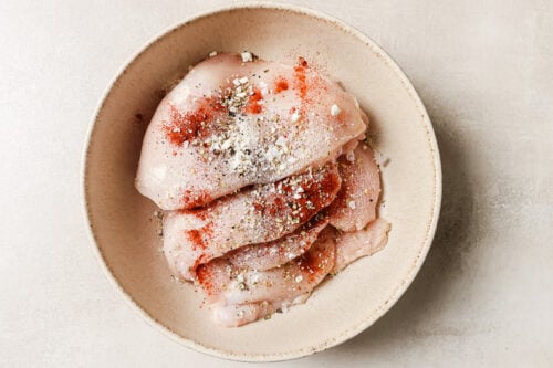 Raw chicken breasts in a beige bowl, sprinkled with seasonings like red paprika and black pepper, ready to be paired with mushrooms for a savory chicken and mushroom dish.