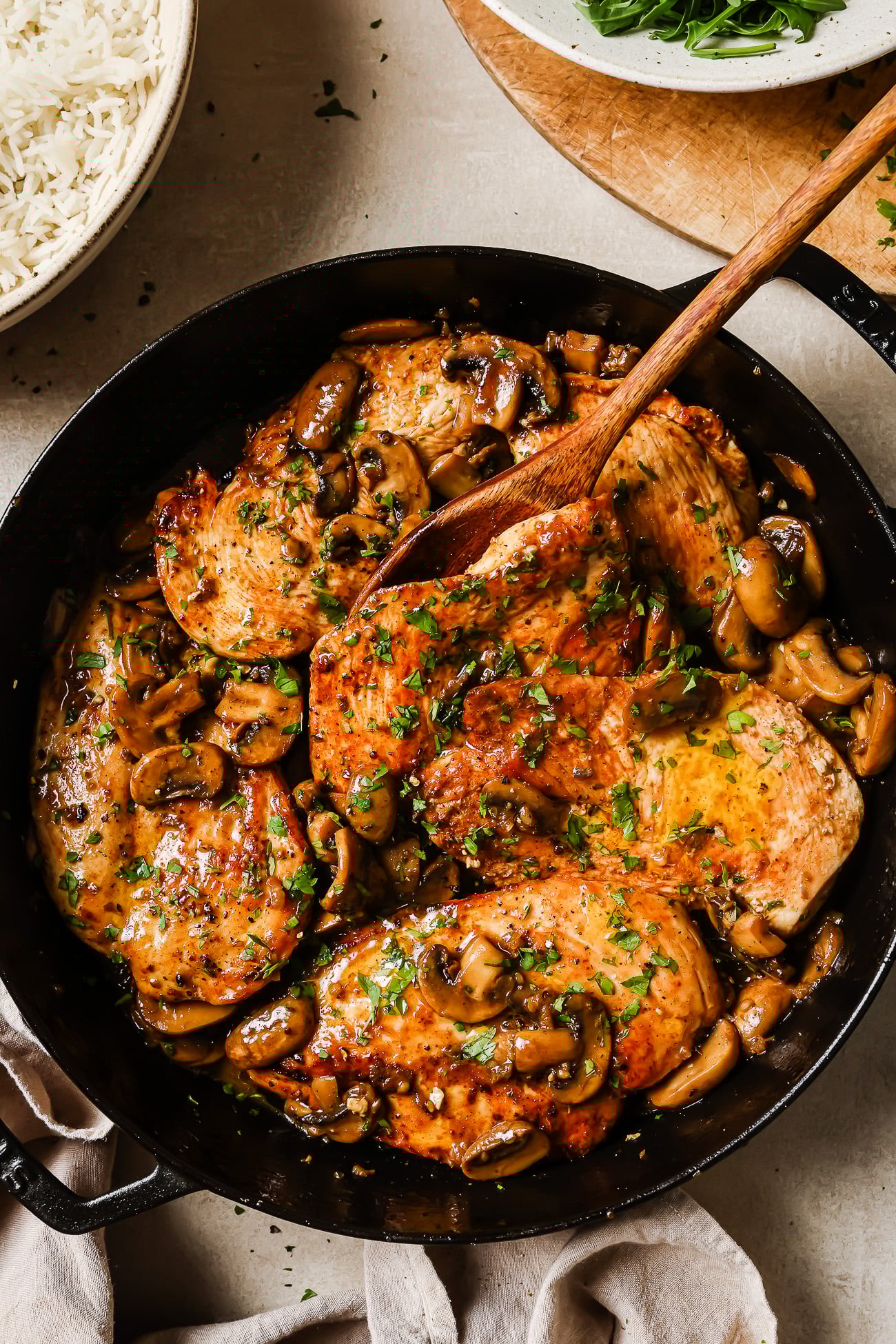 A skillet filled with saut&eacute;ed chicken and mushroom breast in a savory sauce, garnished with chopped parsley, with a wooden spoon serving a piece. Bowls of rice and greens are visible beside the skillet.