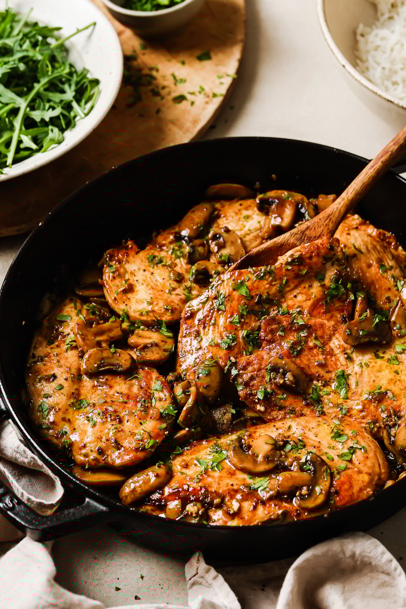 A skillet filled with golden-brown chicken and mushroom breasts, cooked with sliced mushrooms and topped with fresh herbs. A wooden spoon lifts one piece, while fresh greens are visible in the background.