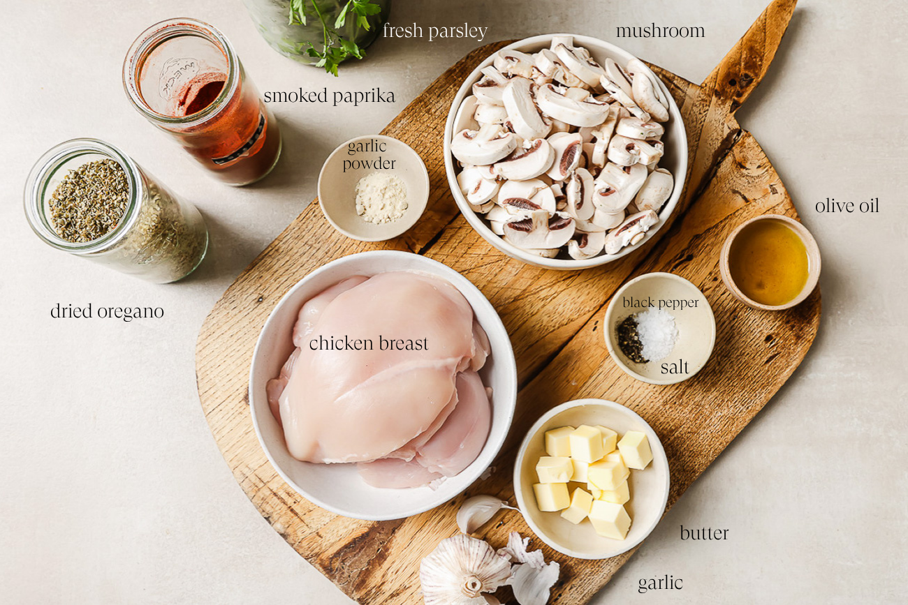 Ingredients for a chicken and mushroom dish on a wooden board: raw chicken breast, sliced mushrooms, butter, garlic, dried oregano, smoked paprika, garlic powder, black pepper, salt, olive oil, fresh parsley, and a glass of red liquid.