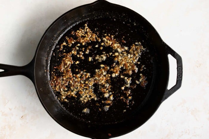 A black cast iron skillet containing browned and saut&eacute;ed minced garlic for a Garlic Butter Shrimp Recipe sits on a light-colored surface.