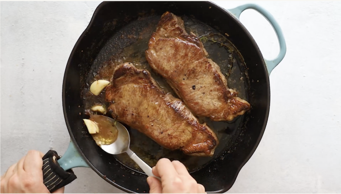 Two browned steaks are cooking in a black cast iron skillet with sprigs of herbs, garlic cloves, and mushrooms. A hand is holding a spoon, scooping up melted butter or juices to baste this delicious pan seared steak with mushrooms.