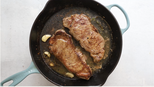 Two seared steaks cook in a cast iron skillet with three cloves of garlic, herbs, and a light brown sauce, ready for the perfect Pan Seared Steak with Mushrooms. The skillet’s blue handle pops against the clean white surface.