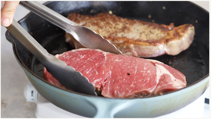 A hand uses metal tongs to place a raw steak into a skillet, while another steak is already searing and browning in the same pan, an essential step for perfect Pan Seared Steak with Mushrooms.