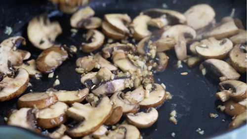 Sliced mushrooms and minced garlic sizzle in a black pan, browning and glistening from the heat—perfect to top a Pan Seared Steak with Mushrooms.
