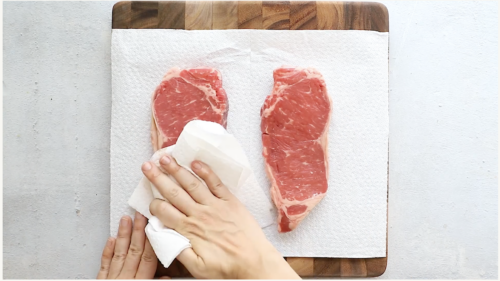 Two raw steaks on a cutting board lined with a paper towel, as a hand pats one steak dry—getting them ready for the perfect Pan Seared Steak with Mushrooms.