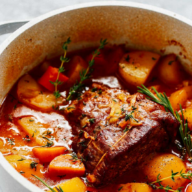 A pot of beef pot roast with potatoes, carrots, and herbs in a rich broth, garnished with rosemary and thyme. Showcasing a classic Beef Pot Roast Recipe, the dish is placed on a gray surface with Beef Pot Roast text above.
