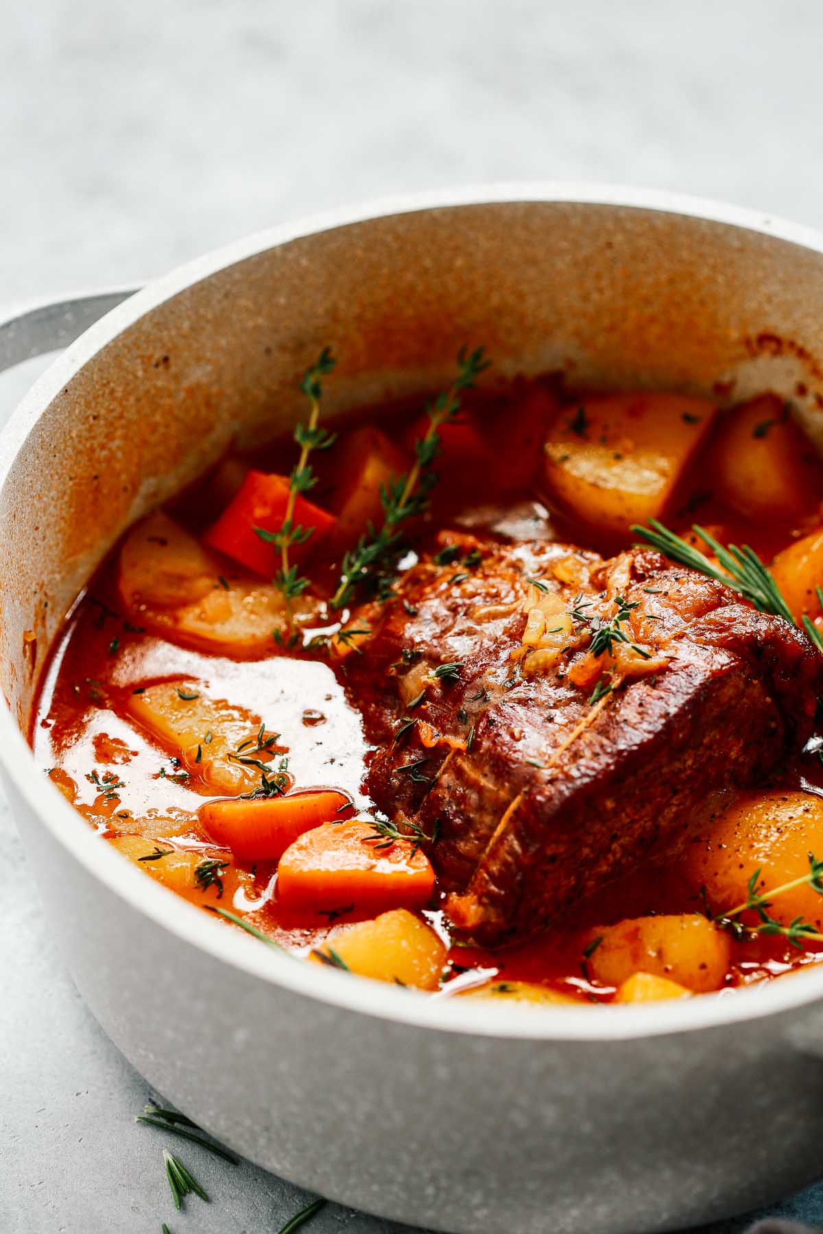 A Beef Pot Roast Recipe simmers in a white pot with carrots, potatoes, and herbs, surrounded by a rich red broth and garnished with fresh thyme and rosemary.