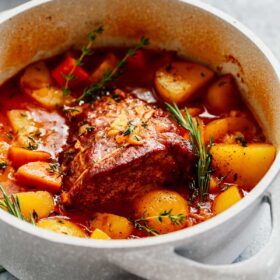 A classic Beef Pot Roast Recipe featuring potatoes, carrots, and herbs simmering in a savory red sauce in a white Dutch oven, sitting on a gray surface with a gray towel nearby.