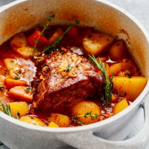 A classic Beef Pot Roast Recipe featuring potatoes, carrots, and herbs simmering in a savory red sauce in a white Dutch oven, sitting on a gray surface with a gray towel nearby.