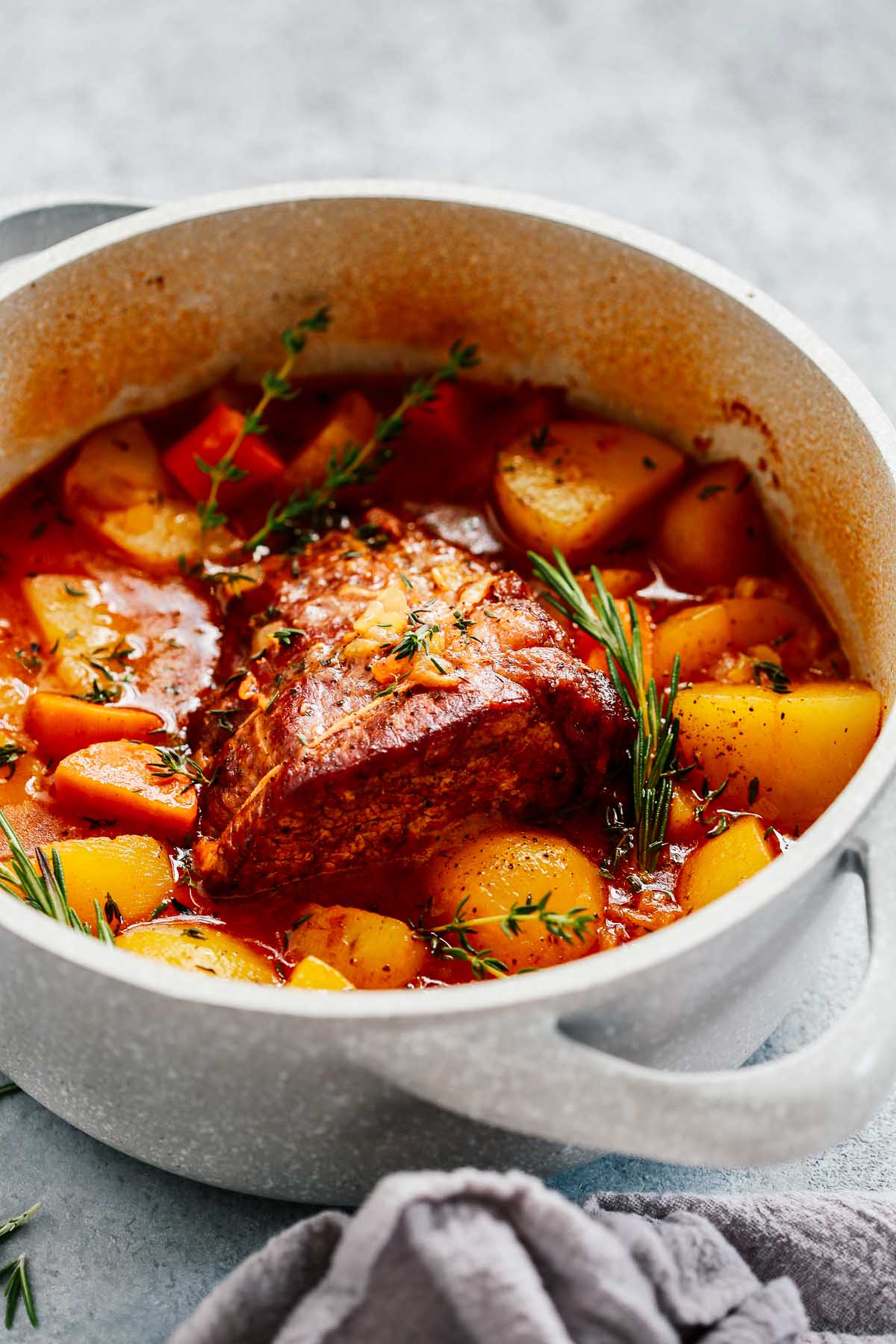 A classic Beef Pot Roast Recipe featuring potatoes, carrots, and herbs simmering in a savory red sauce in a white Dutch oven, sitting on a gray surface with a gray towel nearby.