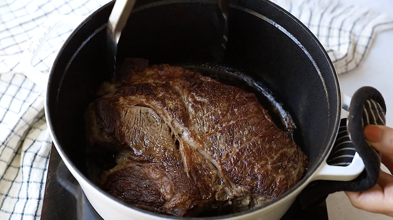 A large piece of browned meat, perfect for a Whole30 Pot Roast, is searing in a black pot, held with metal tongs. A checkered cloth is visible in the background, and a hand is holding the pot’s handle.