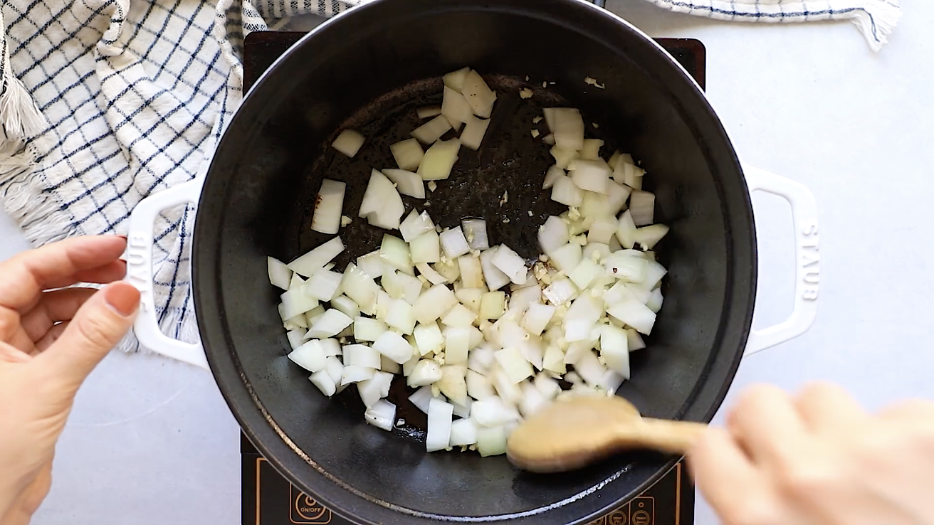Chopped onions being sautéed in a black pot on a stovetop, with a hand stirring them using a wooden spoon—the perfect savory start to your Whole30 Pot Roast. A checkered kitchen towel is visible in the background.