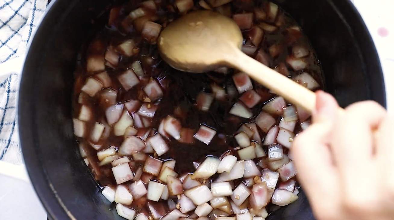 A hand stirs diced onions in a dark liquid with a wooden spoon inside a black pot, likely at the start of cooking a savory Whole30 Pot Roast.