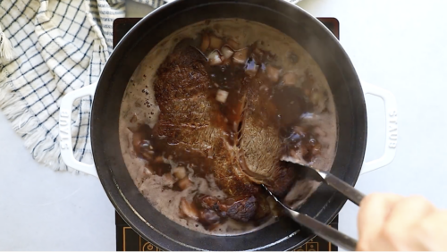 A pair of tongs holds a browned Whole30 Pot Roast in a pot filled with simmering broth and chopped onions on a stovetop, with a kitchen towel nearby.