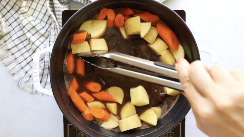 A hand uses metal tongs to stir chopped potatoes and carrots simmering in a dark broth inside a pot on a stovetop as part of preparing a hearty Whole30 Pot Roast. A checkered towel is visible next to the pot.