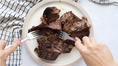 Two hands use forks to shred a cooked piece of Whole30 Pot Roast on a white plate. A checkered towel is partially visible to the left of the plate on a light surface.