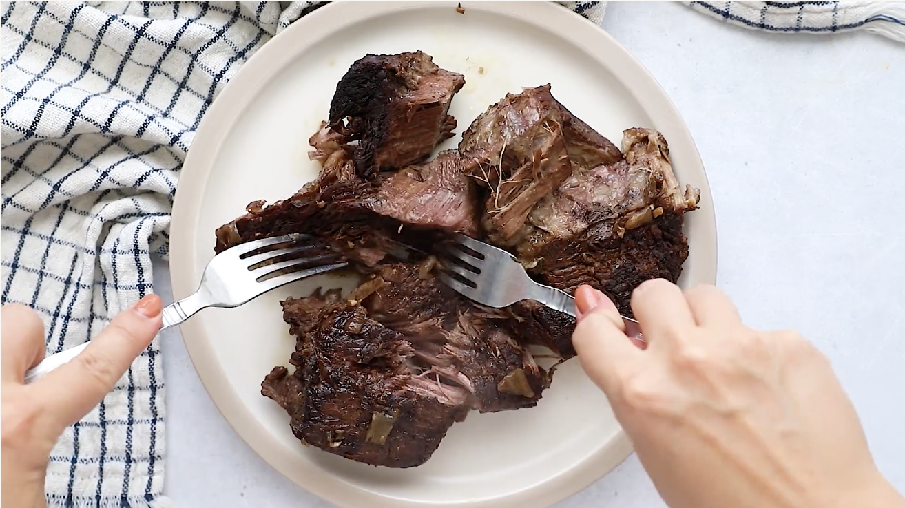 Two hands use forks to shred a cooked piece of Whole30 Pot Roast on a white plate. A checkered towel is partially visible to the left of the plate on a light surface.