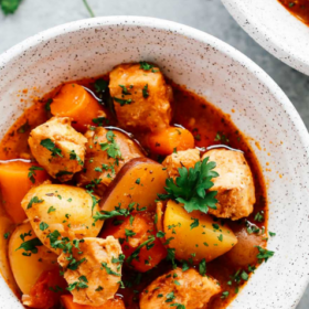 A close-up of a bowl of Instant Pot Chicken Stew with chunks of chicken, potatoes, carrots, and herbs in a tomato-based broth, garnished with parsley. A spoon and another bowl are partially visible.