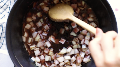 A hand stirs diced onions in a dark liquid with a wooden spoon inside a black pot, preparing the aromatic base for a flavorful Whole30 Pot Roast.