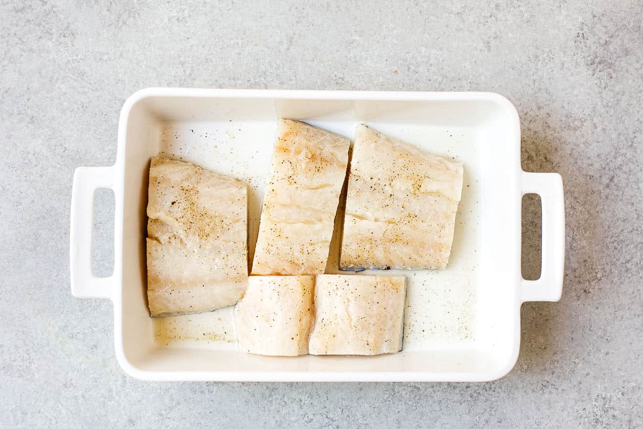 Four raw fish fillets, seasoned with salt and pepper, are placed in a white rectangular baking dish on a light gray textured surface—ideal for preparing a vibrant Brazilian Fish Stew.