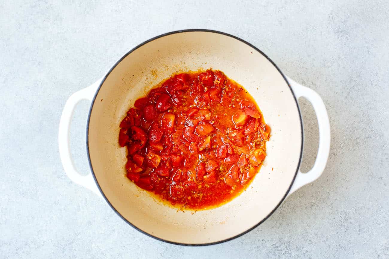 A white pot with handles contains simmering tomato sauce with chunks of tomatoes and herbs, reminiscent of the flavorful base used in Brazilian Fish Stew, on a light gray surface.