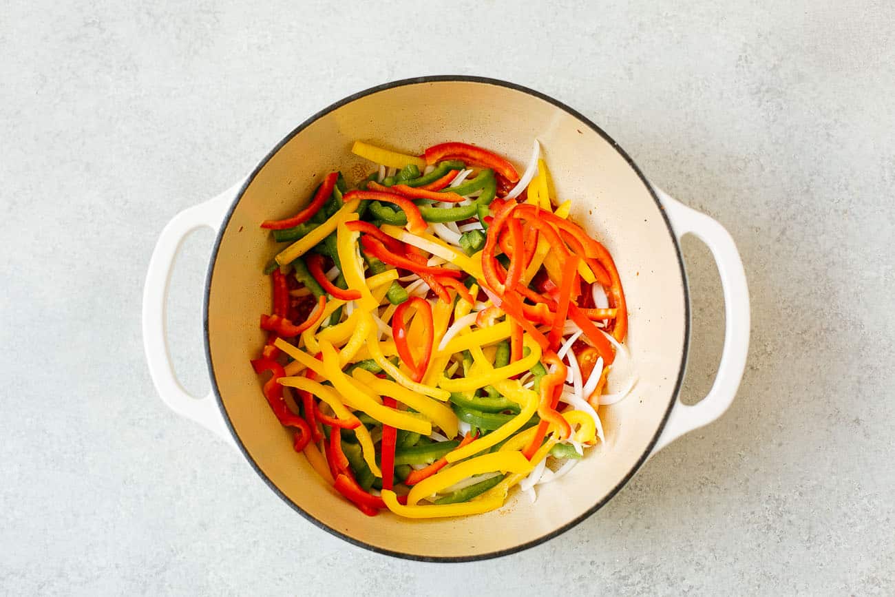 A white pot filled with sliced red, yellow, and green bell peppers and onions, reminiscent of the vibrant ingredients used in a classic Brazilian Fish Stew, placed on a light gray surface.