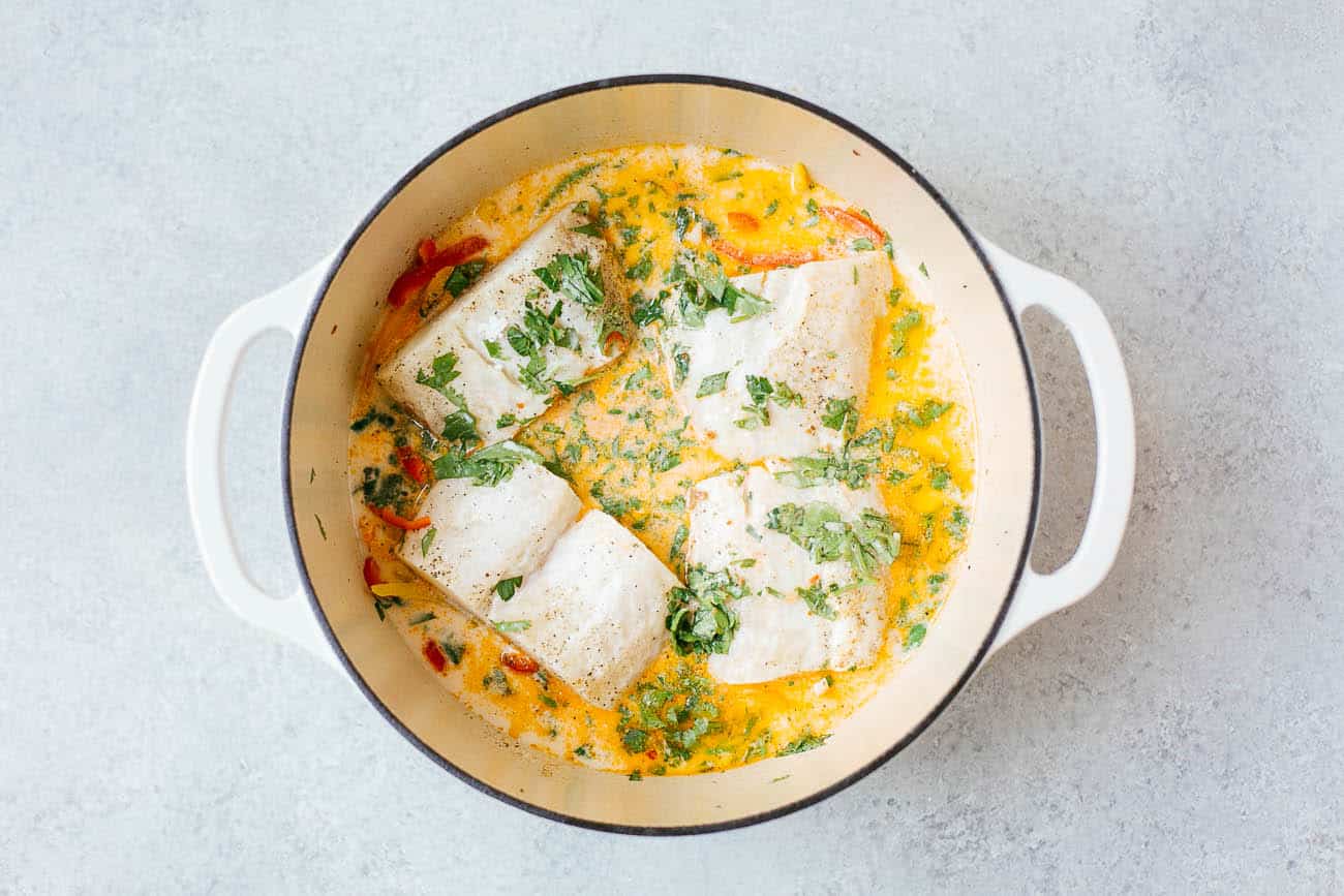 Four pieces of white fish are simmering in a creamy yellow sauce with herbs and vegetables, reminiscent of a classic Brazilian Fish Stew, in a white, round Dutch oven on a light gray surface.